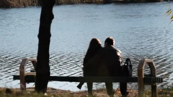 Pareja romántica joven sentada en un banco en un parque de otoño junto al río. Caída. — Vídeos de Stock