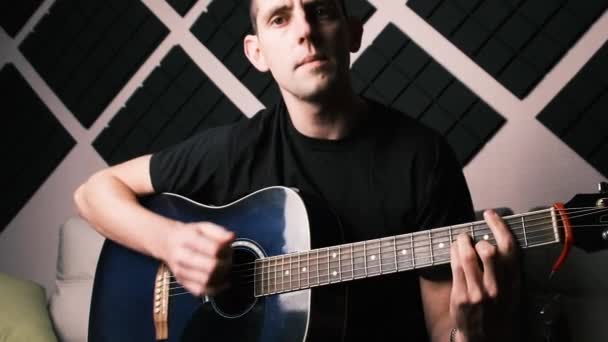 Молодой человек играет на акустической гитаре и поет в домашней студии — стоковое видео