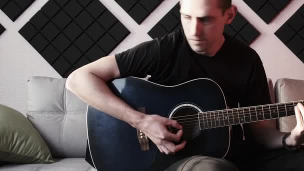 Jovem tocando guitarra acústica enquanto sentado no sofá em estúdio de gravação em casa — Vídeo de Stock