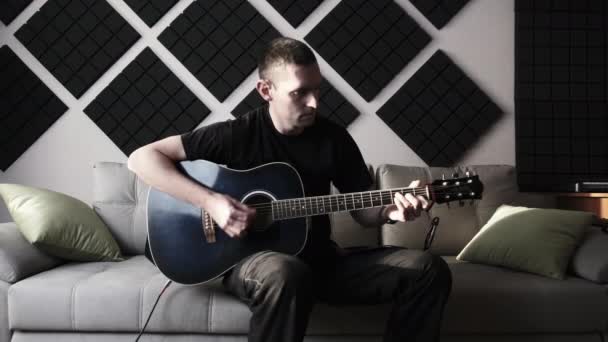 Ung man spelar akustisk gitarr medan du sitter på soffan i hemmet inspelningsstudio — Stockvideo