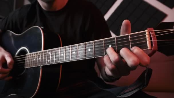 Молодой человек играет на акустической гитаре, сидя на диване в домашней студии звукозаписи — стоковое видео