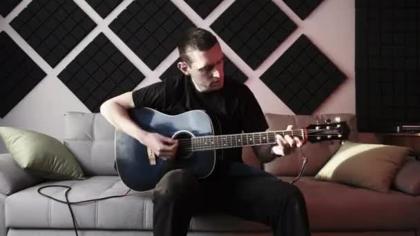 若い男アコースティックギターを演奏しながら自宅録音スタジオでソファに座って — ストック動画