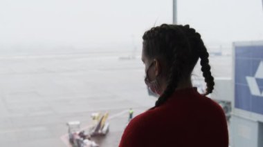 Havaalanında maskeli genç bir kadın gezginin silüeti uçaklara bakıyor.