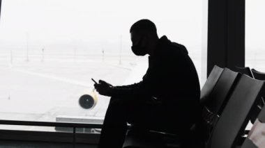 Havalimanı Bekleme Odasında Maskeli Genç Adam Oturuyor ve Pencerenin yanında Akıllı Telefon Kullanıyor
