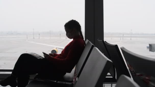 Maskovaná mladá žena v čekárně na letišti sedí a pomocí Smartphone podle okna — Stock video