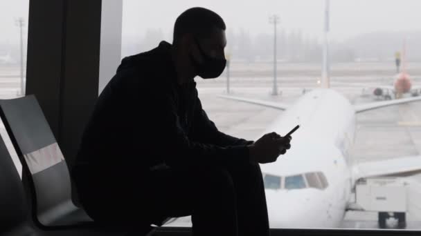 Maskerad ung man på flygplatsen väntar Room Sits och med hjälp av en smartphone vid fönstret — Stockvideo