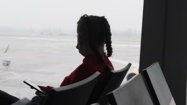Mujer joven enmascarada en la sala de espera del aeropuerto se sienta y usa un teléfono inteligente por la ventana — Vídeo de stock