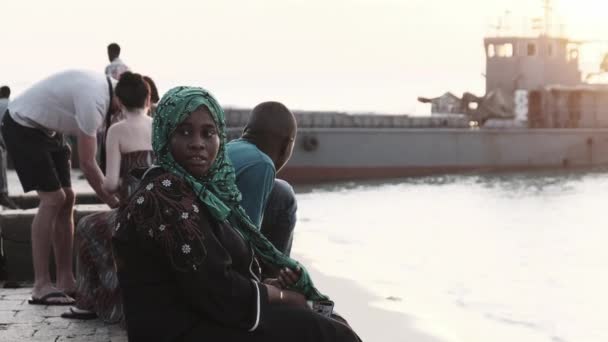 ヒジャーブのアフリカ系イスラム教徒の女性は日没を見るために海の近くの遊歩道に座っている — ストック動画