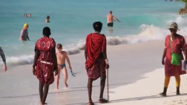 Masai, Tanzanya 'nın Zanzibar kentindeki turistler arasında okyanus kıyısındaki sahil boyunca yürüyor.