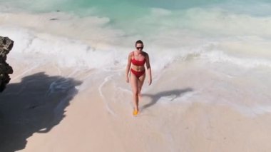 Kırmızı mayo giymiş genç bir kadın Cennet Sahili 'ndeki Turkuaz Okyanusu' na yürür.