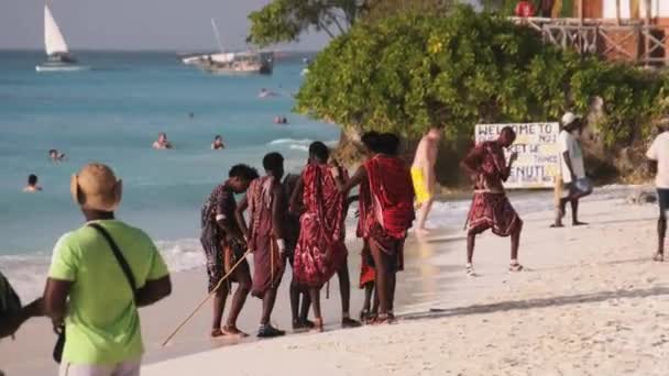 A group of Maasai men stands on the sand beach near Ocean among tourists, Zanzibar, Tanzania. — Stock Video