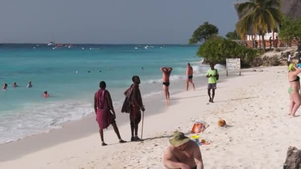 マサイはタンザニアのザンジバルの観光客の間で海の近くのビーチに沿って歩く — ストック動画