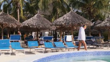 Palmiye ağaçlarının ve Thatched şemsiyelerinin arasında güneşli bir otel ve havuz.