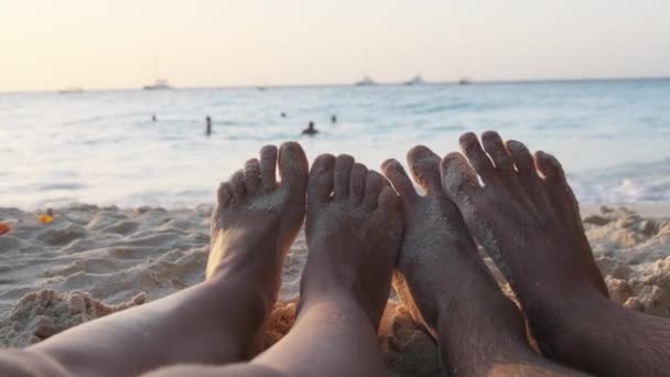 POV Pies de un par de hombres y mujeres acostados en una playa de arena al atardecer por el océano — Vídeo de stock