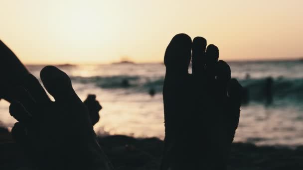 POV Sylwetka stóp młodego mężczyzny leżącego na piaszczystej plaży nad oceanem podczas zachodu słońca — Wideo stockowe