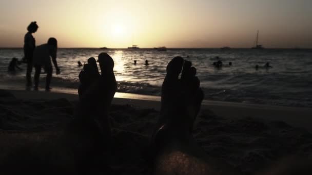 POV silueta de los pies de un joven acostado en la playa de arena por el océano durante la puesta del sol — Vídeo de stock