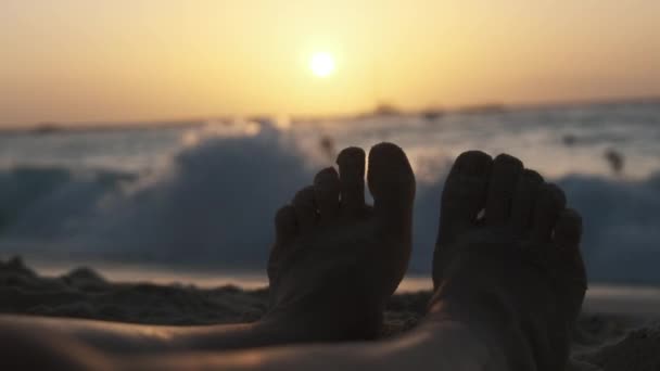 Силуэт ног молодого человека, лежащего на песчаном пляже у океана во время заката — стоковое видео