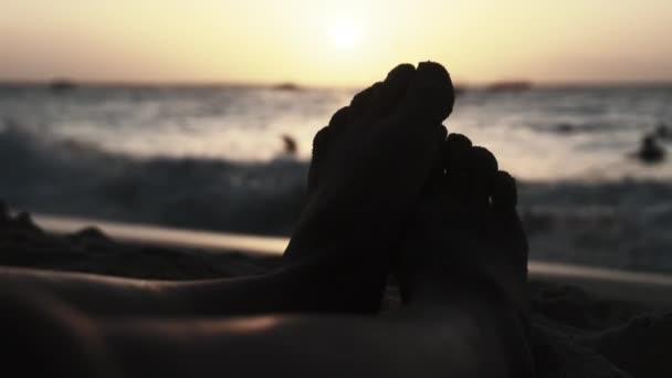 POV silueta de los pies de un joven acostado en la playa de arena por el océano durante la puesta del sol — Vídeos de Stock