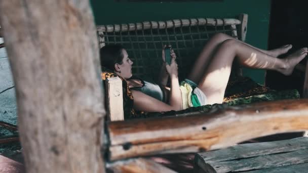 Νεαρό κορίτσι Διαβάστε το βιβλίο από Smartphone που βρίσκεται στην Tropic Veranda της Αφρικής Bungalow — Αρχείο Βίντεο