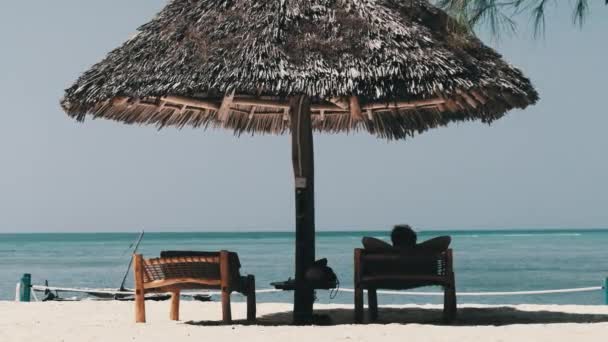 Reetgedeckte Sonnenschirme und Liegestühle mit Mann am Sandstrand am Meer, Sansibar — Stockvideo