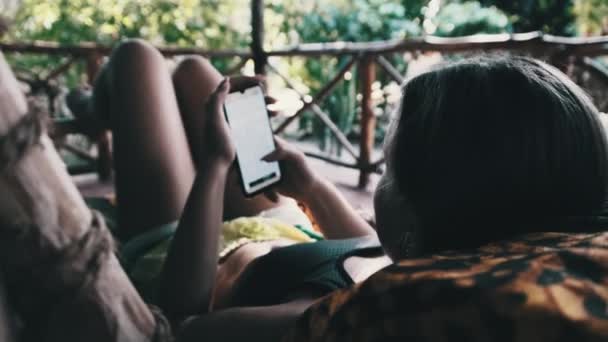 Jovencita Lea el libro desde el teléfono inteligente acostado en la veranda tropical de la cabaña africana — Vídeos de Stock