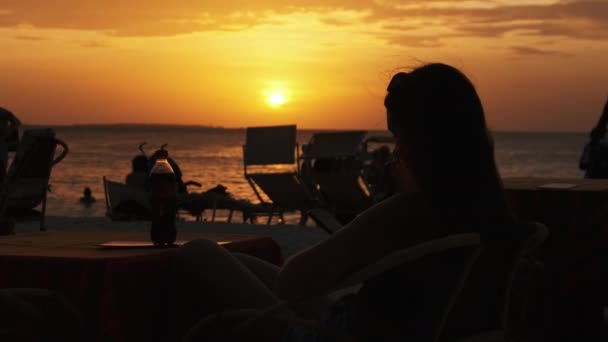 Silhouette Jeune femme assise à table sur la plage et regardant le coucher du soleil par l'océan — Video