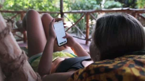 Jovem Leia Livro De Smartphone Deitado na varanda Trópica de Bungalow Africano — Vídeo de Stock