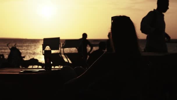 Silhouette Νεαρή γυναίκα κάθεται στο τραπέζι στην παραλία και κοιτάζοντας το ηλιοβασίλεμα από τον ωκεανό — Αρχείο Βίντεο