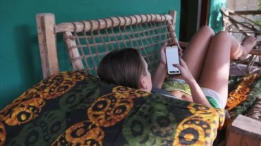 Genç kız Afrika Bungalovunun tropik verandasında uzanan Akıllı Telefondan Kitap Oku