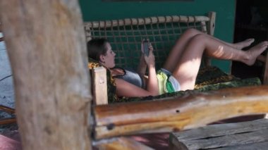 Genç kız Afrika Bungalovunun tropik verandasında uzanan Akıllı Telefondan Kitap Oku