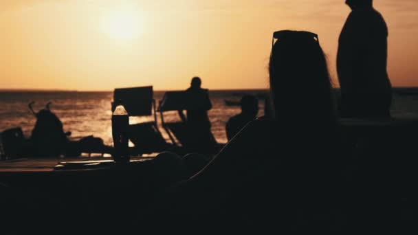 Silhouette Jeune femme assise à table sur la plage et regardant le coucher du soleil par l'océan — Video