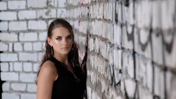 Hermosa joven de pie junto a la pared de ladrillo y mirando a la cámara — Vídeos de Stock