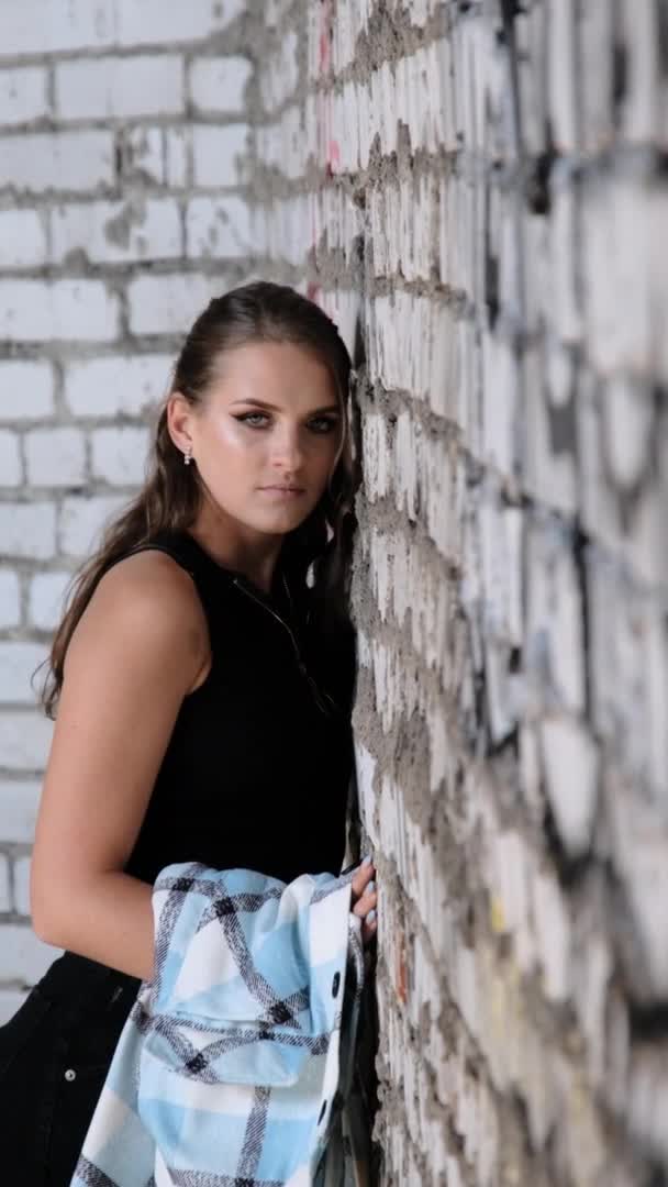 Pionowe wideo Piękna młoda dziewczyna stojąca przy ścianie z cegły i patrząc na aparat — Wideo stockowe