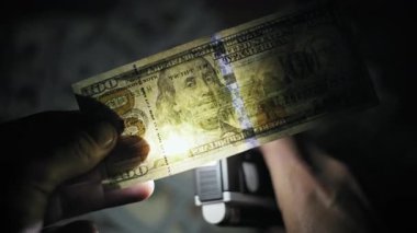 El feneri güvenlik ipliğini, filigran portresini incelerken 100 dolarlık banknot tutuyor. Karanlık, gölgeli ortamda para biriminin kimlik doğrulamasının yakın görüntüsü ve ışık kaynaklı mercek parlaması