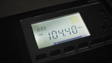 Karanlıkta arka ışıklandırılmış dijital radyo ekranının FM frekanslarını yaklaş. Bir kanalı ararken numaralar hızla dalgalanıyor, frekansın altında bir sinyal gücü ölçer var..