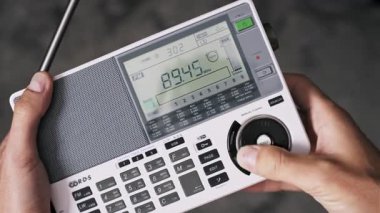 El ele tutuşup taşınabilir beyaz bir dijital radyoyu ayarlarken yüksek açılı bir görüntü. Başparmak, dijital sinyal sayacının yanındaki LCD ekranında değişen FM frekanslarını tarayarak elle arama yapar.