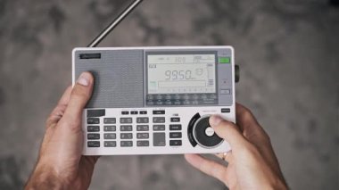 Eller beyaz portatif telsizi karanlık yüzey üzerinde tutar, FM bandını taramak için elle çevirir. Arka plandaki dijital ekran frekans artışı gösteriyor, anten ile açık yayın sinyali aranıyor.