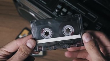 Boş beyaz etiketli, şeffaf kabuklu, klasik ses kasedini ters çeviren ellerin makro görüntüsü. Makara ve B tarafında kırmızı ayrıntı gösteriliyor, yumuşak odak arkaplanındaki retro teyp oynatıcısı ile birlikte.