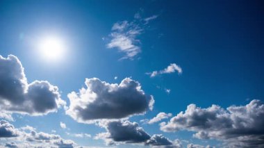 Hızlı hareket eden beyaz ve gri bulutlar derin mavi gökyüzünde yüzüyor, parlak güneşi geçiyor, berrak gökyüzünün parçalarını açığa çıkarıyor ve güneşli bir günde dinamik gölgeler bırakıyor..