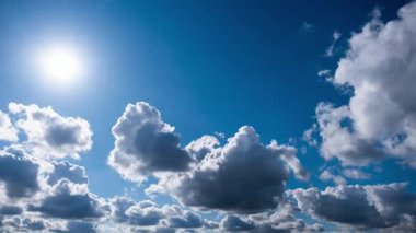 Derin mavi gökyüzünde ilerleyen beyaz kümülüs bulutlarının hızlandırılmış görüntüsü. Parlak güneş, daha karanlık fırtına bulutlarının oluşmaya başlamasıyla parlıyor. Hava güneşli, bulutlu ve dramatik bir sahne yaratıyor..