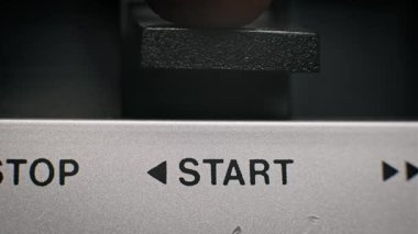 Bir başparmağın gümüş fırçalı metal konsoldaki siyah dikdörtgen durdurma düğmesine basmasının makro perspektifi. Yazdırılmış tipografiyi temizle bir retro ses veya video makinesini çalıştırmak için işlevi tanımlar.