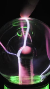 Bir parmağın plazma küre yüzeyine dokunuşu. Pembe ve yeşil elektrikli civatalar birleşir ve parmak ucunu takip eder. Merkezi elektrot koyu arkaplanda görünür. Etkileşimli yüksek voltaj deneyi.