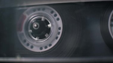 Klasik bir ses kasetinin yakın çekim görüntüsü bir çalgıda geri sarılıyor. Dairesel delikleri olan beyaz plastik makara, şeffaf kaplamanın içindeki makaranın manyetik bandını sarar..