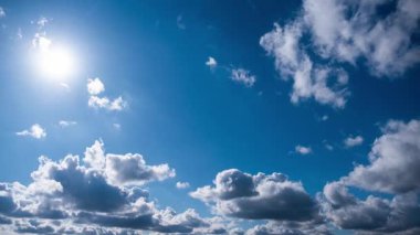 Derin mavi gökyüzünde ilerleyen beyaz ve gri kümülüs bulutlarının hızlandırılmış görüntüsü. Sol üst köşedeki parlak güneş ışık saçar ve açık, güneşli bir günde doğal bir lens patlaması yaratır..