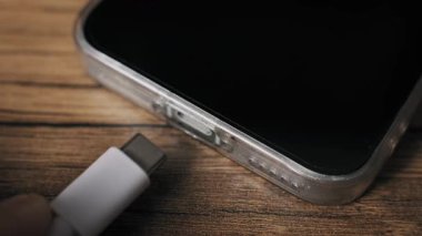 Akıllı telefon şarj portuna beyaz bir USB-C kablosu sokan bir elin makro görüntüsü. Cihazın şeffaf bir çantası ve ahşap bir masanın üzerinde duran siyah bir ekranı var. Güç için bir aygıt bağlanıyor.