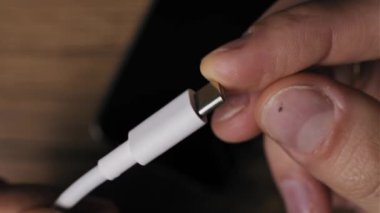 El beyaz bir USB-C kablosu tutuyor, konnektör ucuna odaklanıyor, sonra da akıllı telefona takıyor. Bulanık ekran, dairesel şarj yüzdesi simgesi ile aydınlanıyor. Modern güç dizisi.