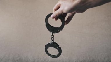 Siyah metal kelepçelerle basit bir arkaplanda sallanan bir erkek eli. Zincir ve kilit mekanizması, ceza adaleti, tutuklama ve kolluk kuvvetlerini sembolize ediyor..