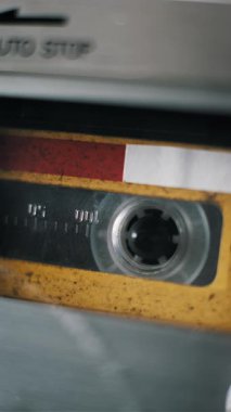 Klasik ses kasetlerinin dikey, aşırı yakın plan görüntüsü şeffaf makaralar çalınırken dönüyor. Sarı ve kırmızı kaset etiketi, kayıt cihazındaki manyetik kaset ilerledikçe görünür hale geliyor..