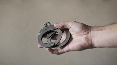 Tarafsız bir arkaplanda metal bir kelepçeyi tutan elin yakın plan çekimi. Kişi kelepçeleri sarkıtır ve döndürür. Zincirleme ve kilitleme mekanizmasını gösterir..
