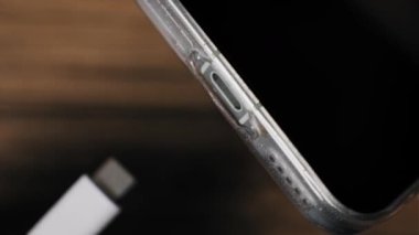Bir elin akıllı telefon yan portuna beyaz bir C tipi konnektör yerleştirme açılı makro perspektifi. Temiz bir tampon kutusundaki hücresel cihaz, karanlık bir ahşap arka planda elektrik akımı alıyor..
